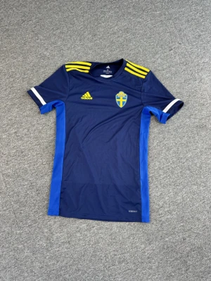 Blå Sverige Adidas matchtröja - Säljer en blå matchtröja från Adidas med Sveriges landslagsemblem på bröstet. Tröjan har gula detaljer på axlarna, vita ränder på ärmarna och blå sidopaneler. Tillverkad i ett lätt och ventilerande material, perfekt för träning eller match.