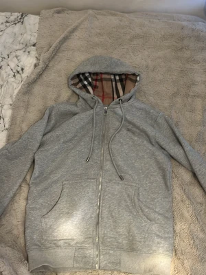Grå zip hoodie med Burberry-mönster - Snygg grå zip hoodie från Burberry med klassiskt rutigt foder i huvan. Hoodien har dragsko i huvan, två fickor fram och ribbade muddar. Perfekt för dig som vill ha en stilren men ändå lyxig look. Tillverkad i mjuk bomullsblandning.