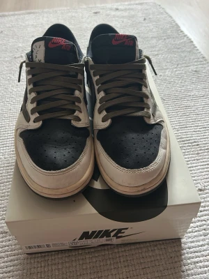 Riktiga  Jordan 1 Low Travis Scott - Rikgita  Jordan 1 Low Travis Scott med svart tåbox och paneler, vita overlays och olivgröna skosnören. Swoosh-loggan är brun och vänd åt fel håll på utsidan. Röd Nike Air-logga på plösen. Materialmix av läder och mocka för en unik look.