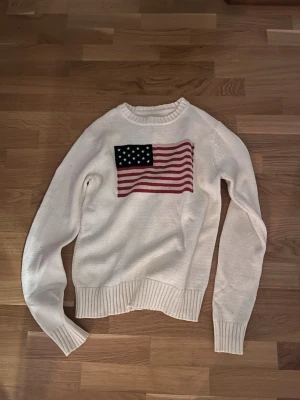 Vit stickad tröja med USA-flagga - Vit stickad tröja med rund hals och ribbade muddar. På bröstet finns en stor amerikansk flagga i rött, vitt och blått. Tröjan har en avslappnad passform och är perfekt för dig som gillar klassisk och preppy stil.