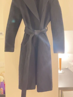 Svart kappa från Vero Moda - Stilren svart kappa från Vero Moda med knytskärp i midjan och klassisk krage. Lång modell som passar perfekt till kyligare dagar. Enkel och tidlös design som är lätt att matcha med olika outfits.