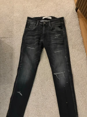 Svarta slim fit jeans med slitningar - Snygga svarta slim fit jeans från Replay med slitna detaljer och diskreta färgstänk. Jeansen har klassisk femficksdesign, dragkedjegylf och smal passform. Perfekta för en trendig och avslappnad look.