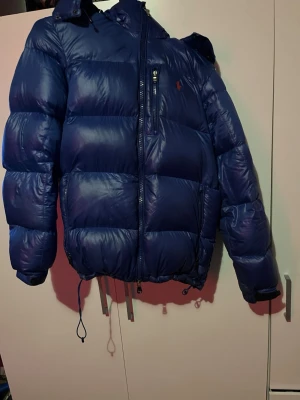 Blå dunjacka från Polo Ralph Lauren - Snygg blå dunjacka från Polo Ralph Lauren med huva, dragkedjor framtill och på bröstfickan samt justerbar dragsko i nederkant. Jackan är rejält vadderad och perfekt för kalla vinterdagar. Klassisk puffer-modell med diskret logga på bröstet.