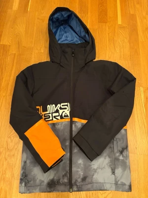 Quiksilver skidjacka/vinterjacka med huva - Snygg skid/vinterjacka från Quiksilver i svart, grått och orange. Jackan har huva, dragkedja framtill och tryck på bröstet. Finns även liftkortsficka på ärmen. Fint skick. Inte mycket använd. Storlek w14 men skulle säga som 164/170/xs.