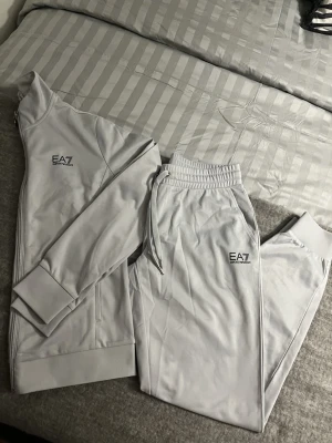 Grått loungewear-set från EA7 - Mysigt grått loungewear-set från EA7 Emporio Armani med zip-jacka och matchande byxor. Setet har stilren logga på både jacka och byxa, ribbade muddar och dragkedja framtill. Materialet är mjukt och stretchigt, perfekt för chill hemma eller avslappnade dagar.