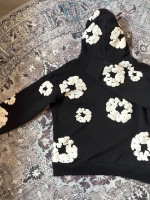 Denim tears set - Svart hoodie med stora vita blommönster över hela tröjan. Tröjan har huva och en klassisk känguruficka framtill. Långärmad och i mjukt bomullsmaterial, perfekt för en chill och trendig look. Medföljer shorts