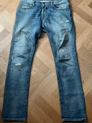 Blå slitna jeans med raka ben - Säljer ett par blå jeans med raka ben och slitna detaljer på både fram- och baksida. Jeansen har klassisk femficksdesign, knappgylf och är tillverkade i denim. Perfekta för en avslappnad och trendig look.