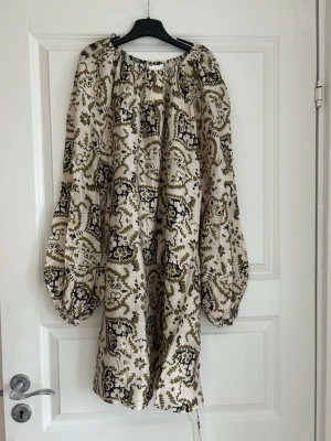 Mönstrad kortklänning med volymärm - Luftig kortklänning med beige botten och grönt mönster. Klänningen har långa volymärmar, omlottknytning i midjan och avslappnad passform. Perfekt för dig som gillar bohemisk stil och unika detaljer.