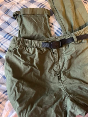 Olivgröna chinos med bälte - Snygga olivgröna chinos med elastisk midja och justerbart svart bälte. Byxorna har raka ben och är tillverkade i ett mjukt bomullsmaterial. Perfekta för en avslappnad och stilren look. Byxorna är lite nötta på knäna/lår och på baksida .