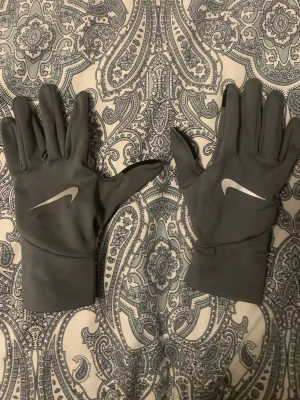 Grå Nike löparhandskar - Snygga grå löparhandskar från Nike med reflekterande logga på ovansidan. Handskarna är tunna och smidiga, perfekta för utomhusträning när det är kallare. De har en stilren design och sitter tajt runt handleden.