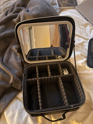 Sminkspegel med LED och förvaring - Praktisk sminkspegel med inbyggd LED-belysning och touch-knapp. Spegeln sitter i locket på en svart, hård sminkväska med justerbara fack och elastiska fickor för sminkborstar och tillbehör. Perfekt att ta med tack vare bärhandtaget och robust design. Finns olika inställningar på spegelljuset. Belysningen laddas via USB-C. Mått: 23,4 x 26 x 10 cm. Nypris 499kr.