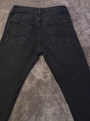 Svarta jeans från Jack & Jones - Snygga svarta jeans från Jack & Jones med klassisk femficksdesign och raka ben. Jeansen har en diskret tvättad look och är tillverkade i ett mjukt denimtyg med stretch för extra komfort. Perfekta för en stilren och avslappnad outfit.