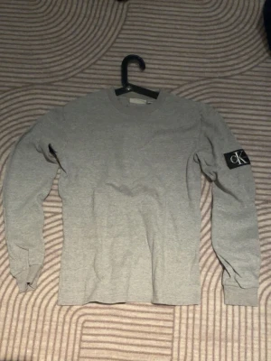 Grå långärmad tröja från Calvin Klein - Snygg grå långärmad tröja från Calvin Klein med rund halsringning och en svart patch med CK-logga på ena ärmen. Tillverkad i mjukt bomullsmaterial som känns skönt mot huden. Enkel och stilren design som passar till det mesta.