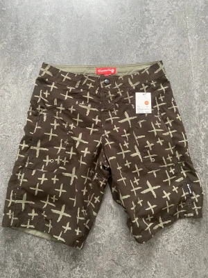 Bruna mönstrade shorts från Superdry - Coola bruna shorts från Superdry med unikt ljusgrönt korsmönster. Shortsen har flera fickor framtill och bak samt knapp- och dragkedjestängning. Perfekta för sommardagar när du vill sticka ut lite extra. Är gjorda för bad till barn . Köpta för 300kr 