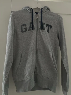 Grå zip-hoodie från GANT - Snygg grå hoodie från GANT med dragkedja framtill och mörkblå snören i huvan. Stor GANT-logga i mörkblått på bröstet och två fickor framtill. Mjuk bomullsblandning och klassisk passform, perfekt för en avslappnad stil.