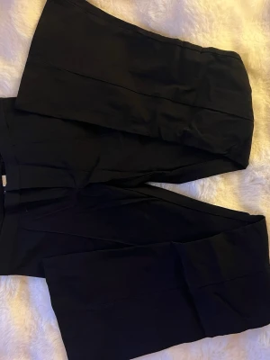 Svarta vida kostymbyxor - Snygga svarta kostymbyxor med vida ben och pressveck. Byxorna har en klassisk passform och är perfekta för en stilren look. Materialet är ett mjukt tyg med lite struktur, vilket ger en dressad känsla. I storlek 36/34💗