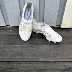 Adidas Predator vita fotbollsskor - Säljer ett par Adidas Predator fotbollsskor i vitt med blå och silverdetaljer. Skorna har snörning, klassisk design och är tillverkade i syntetmaterial med texten HYBRIDFEEL på sidan. Perfekta för dig som vill sticka ut på planen.