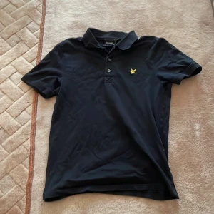 Svart pikétröja från Lyle & Scott M - Klassisk svart pikétröja från Lyle & Scott i storlek M. Tröjan har korta ärmar, krage och knäppning framtill med tre knappar. På bröstet sitter den ikoniska gula fågel-loggan. Perfekt för en clean och stilren look.     PRIS DISKUTERAS! DU SOM KÖPER BETALAR OCKSÅ FRAKTEN!