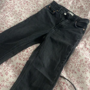 Svarta gina jeans med rå kant - Säljer ett par svarta Gina jeans med klassisk femficksdesign och knappgylf. Jeansen har en rak passform och rå, fransig kant längst ner på benen för en cool och avslappnad vibe. Perfekta till sneakers eller boots.