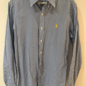 Randig blå skjorta från Polo Ralph Lauren - Snygg blå och vit randig skjorta från Polo Ralph Lauren i slim fit. Klassisk krage, långärmad och med knappar hela vägen framtill. Ikonisk gul broderad logga på bröstet. Tillverkad i mjuk bomull som känns skön mot huden.