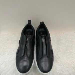 Valentino Garavani svarta sneakers - Svarta sneakers från Valentino Garavani i skinn med vita sulor dekorerade med små nitar runtom. Snygg minimalistisk design utan snörning och med rund tå. Perfekta för dig som gillar lyxiga och stilrena skor med en edgy touch.