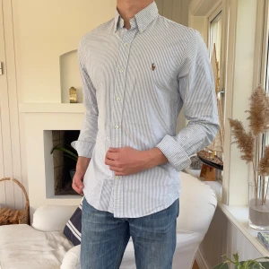 Randig skjorta från Ralph Lauren M - Snygg slim fit skjorta från Ralph Lauren i vitt och ljusblått med tunna ränder. Klassisk button-down krage, broderad logga på bröstet och långa ärmar. Skjortan är i bomull och har knappar framtill. Perfekt för en clean och stilren look.