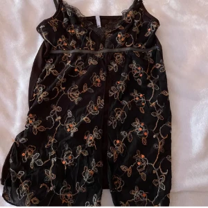 Svart blommig spetsklänning från Miss Selfridge - Svart kortklänning med tunna axelband och vacker spetskant vid urringningen. Framsidan är dekorerad med broderade blommor i beige, orange och guldtoner. Klänningen har en feminin passform och är perfekt för dig som gillar detaljer och blommigt.