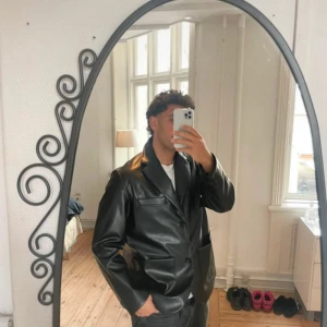 Svart skinnliknande jacka/kavaj med klassisk krage - Säljer en svart skinn liknande kavaj/jacka med klassisk krage och knäppning framtill. Jackan har en stilren look med raka linjer, två stora fickor och en bröstficka. Passar dig som vill ha en edgy och modern vibe.