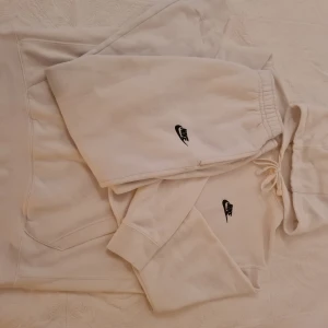 Vit Nike mjukisdress med broderad logga - Säljer en fräsch vit Nike mjukisdress  i mjuk bomull med elastisk midja och snörning. Klassisk svart Nike-logga broderad framtill. Perfekta för chill dagar eller träning. Snygg och enkel design med fickor på sidorna.