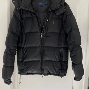 Svart dunjacka Polo Ralph Lauren - Svart dunjacka från Polo Ralph Lauren med röd broderad logga på bröstet. Jackan har några mindre lagningar, men de syns knappt. Modellen heter The Gorham Down Jacket och kostar 5795kr på Ralph Laurens hemsida. Skriv vid frågor:)