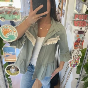 Grön overshirt med volang och fransar - Säljer en ljusgrön overshirt med dragkedja och volangdetalj nertill. Jackan har långa ärmar med rynkade muddar och dekorativa fransar i vitt längs bröstet. Perfekt för dig som gillar en chill och bohemisk stil.