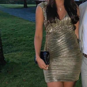 Skimrig grön klänning från Maje - Supersnygg kort klänning i skimrigt grön tyg från Maje. Plisserad design med omlottkänsla och djup v-ringning. Ärmlös modell som sitter tight och markerar midjan. Perfekt för dig som vill sticka ut med metallic vibes och cool struktur.