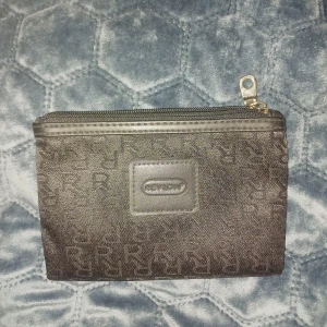 Svart plånbok från Revlon med dragkedja - Snygg svart plånbok från Revlon med diskret mönster av R-bokstäver över hela ytan. Praktisk dragkedja upptill i metall och stilren, rektangulär form. Perfekt för att hålla koll på kort och småsaker. Den är svartare i verkligheten. Skriv privat för flera frågor 