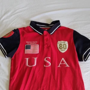 Röd pikétröja chief keef - Röd pikétröja med marinblå krage och ärmar, vita ränder på krage och ärmslut. Stort USA-tryck framtill, broderad amerikansk flagga