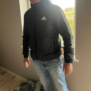 Svart pufferjacka från Adidas M - Svart pufferjacka från Adidas med huva och klassiska vita ränder längs ärmarna. Jackan har dragkedja framtill, två fickor och Adidas-logga på bröstet. Perfekt för kyliga dagar och har en sportig vibe.