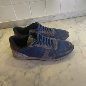 Valentino Garavani blå sneakers - Snygga blå sneakers från Valentino Garavani med detaljer i mocka och skinn. Skorna har klassisk snörning, meshpaneler och en cool kontrast mellan mörkblått och grått. Perfekta för dig som vill ha en exklusiv touch på din streetstyle.