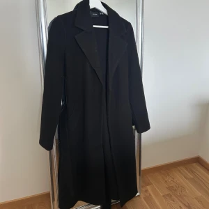 Svart lång kappa från Vero Moda - Svart lång kappa från Vero Moda med klassisk krage och bälte i midjan. Snygg och stilren design med raka linjer och diskreta fickor på sidorna. Perfekt för dig som gillar minimalistisk och clean look. Materialet är mjukt och känns skönt mot kroppen.