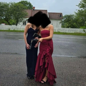 Vinröd långklänning med volang - Säljer en vinröd axelbandslös långklänning med figurnära passform och volangdetaljer längs slitsen framtill. Klänningen har en snygg slits och är tillverkad i ett material som formar sig efter kroppen. Perfekt för bal eller festkvällar! Har använt klänningen en gång så nästan i nytt skick💗