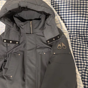 Grå dunjacka Moose Knuckles L - ÄKTA!! Nypris cirka 15k. Säljer en grå dunjacka från Moose Knuckles i storlek L. Jackan har huva, flera fickor med tryckknappar, detaljer med nitar och dragkedja framtill. Klassisk logga på ärmen och insidan. Perfekt för kalla dagar och riktigt snygg streetstil.