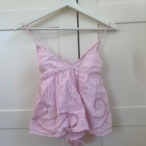 Ljusrosa linnetopp med spetsdetaljer - Söt ljusrosa linnetopp med tunna axelband och v-ringning. Toppen har broderade spetsdetaljer och genombrutna mönster längs fållen. Luftig och flowy passform, perfekt för varma dagar.