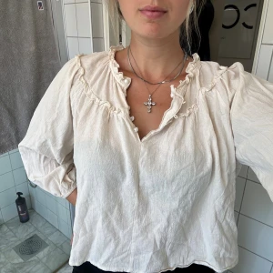 Beige blus med volang och pärlknappar - Supersöt beige blus från Classic Fashion med volangdetaljer längs ringningen och pärlknappar framtill. Blusen har lång ärm med resår vid ärmslut och en lätt, luftig känsla. Perfekt för dig som gillar romantiska och feminina detaljer.