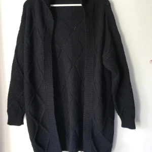 Svart stickad kofta med strukturmönster - Mysig svart stickad kofta med lång ärm och öppet framtill. Koftan har ett diskret strukturmönster i form av stora rutor och bred ribbad kant längs öppningen. Perfekt att slänga på sig när det är kyligt ute. Storlek M/L