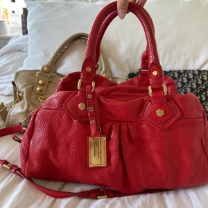 Röd Q Classic Boston Bag  - Perfekta skolväskan! Rymlig röd Q Classic Boston bag från Marc by Marc Jacobs i äkta läder med guldfärgade detaljer och dubbla handtag. Långa bandet medföljer. Klassisk och tidlös modell som rymmer allt du behöver och passar perfekt både till vardag och resa. Praktiska fack och dragkedja gör den enkel att organisera. Välbevarad vintage-modell, men slitage finns. Se alla bilder.  Mått 40x22x15 (undersida). Kan hämtas i Nacka eller skickas. Skriv privat för fler bilder 🤍