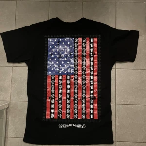  Chrome Hearts USA flagga shirt - Svart t-shirt från Chrome Hearts i bomull med stor amerikansk flagga på ryggen och logga på bröstfickan. Snygga detaljer med vita och röda tryck samt unika Chrome Hearts-mönster i flaggan. Klassisk passform och korta ärmar.