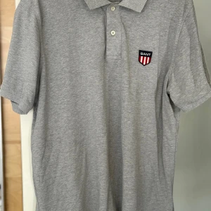 Grå piké från GANT i XL - Snygg grå pikétröja från GANT med klassisk krage, två knappar och broderad GANT-logo på bröstet. Tillverkad i mjuk bomull som känns skön mot huden. Perfekt för en clean och stilren look. SOM NY