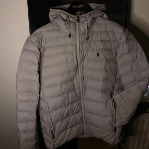 Polo Ralph Lauren jacka - Säljer en riktigt snygg ljusgrå dunjacka från Polo Ralph Lauren. Jackan har en stilren design med loggan broderat på bröstet. Den är använd 2 gånger så nästintill oanvänd. Hör av dig vid fler frågor eller funderingar!