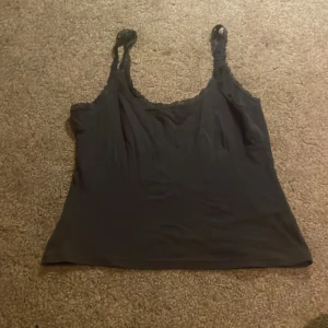 Mörkblå linnetopp med spetsdetaljer - Snygg mörkblå linnetopp med tunna axelband och spetsdetaljer längs ringningen och banden. Toppens material känns mjukt och lätt, perfekt för varma dagar. Enkel och stilren design som är lätt att matcha med olika outfits.