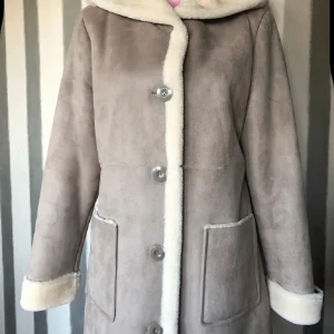 Beige pälsjacka med huva från Junge - Snygg beige pälsjacka från Junge med mysig huva och stora fickor framtill. Jackan har vita pälsdetaljer längs knäppningen och ärmarna, samt stängs med knappar. Perfekt för kalla dagar när du vill vara både varm och stilren. Ej äkta päls 