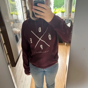 Vinröd hoodie från Dope med tryck - Säljer en vinröd hoodie från Dope med stort vitt tryck på framsidan. Tröjan har huva med snörning, magficka och långa ärmar. Materialet är mjuk bomull och passformen är normal. Perfekt för chill dagar och streetstyle. En blekt fläck bakom armbågen men syns inte tydligt när man har den på