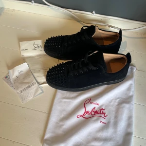 svarta sneakers med nitar - Svarta sneakers i mocka med coola svarta nitar på tån och klassisk röd sula med logga. Skorna har snörning och en platt sula, perfekt för dig som gillar edgy designerstil. 
